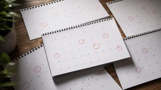Uma visão de cima de várias páginas de um calendário de papel, mostrando semanas que passam, com alguns dias circulados e outros em branco, simbolizando o planejamento e a passagem do tempo real para concluir um curso.